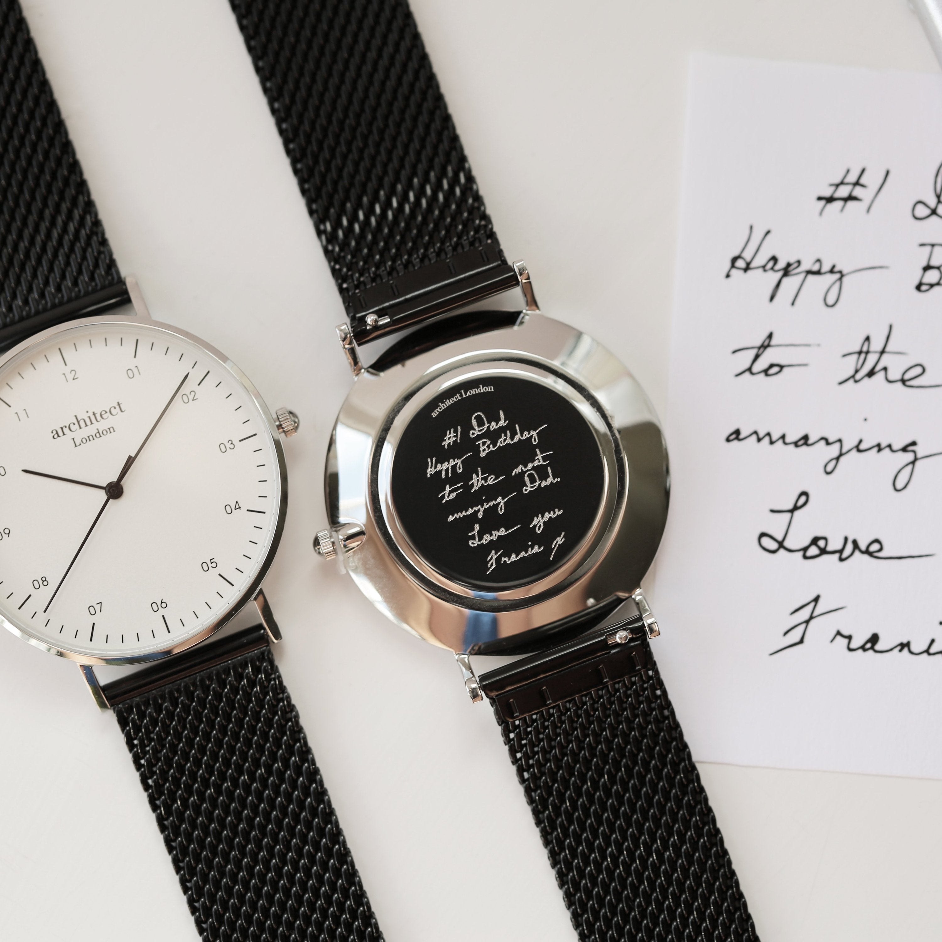 Two watches with black mesh straps on a white surface with handwritten text.
