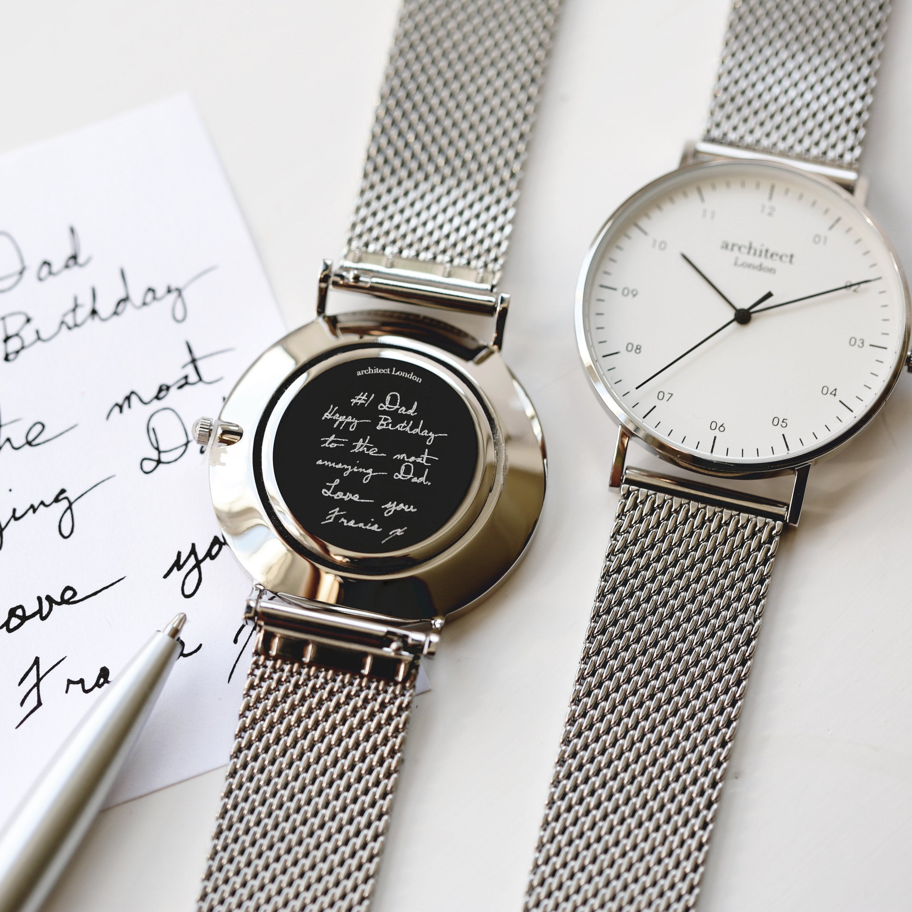 Two silver watches with mesh straps on a white surface with handwritten text.