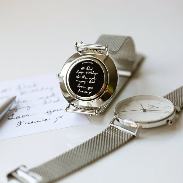 Silver watch with a message on the face, placed on a white surface with a pen and paper.