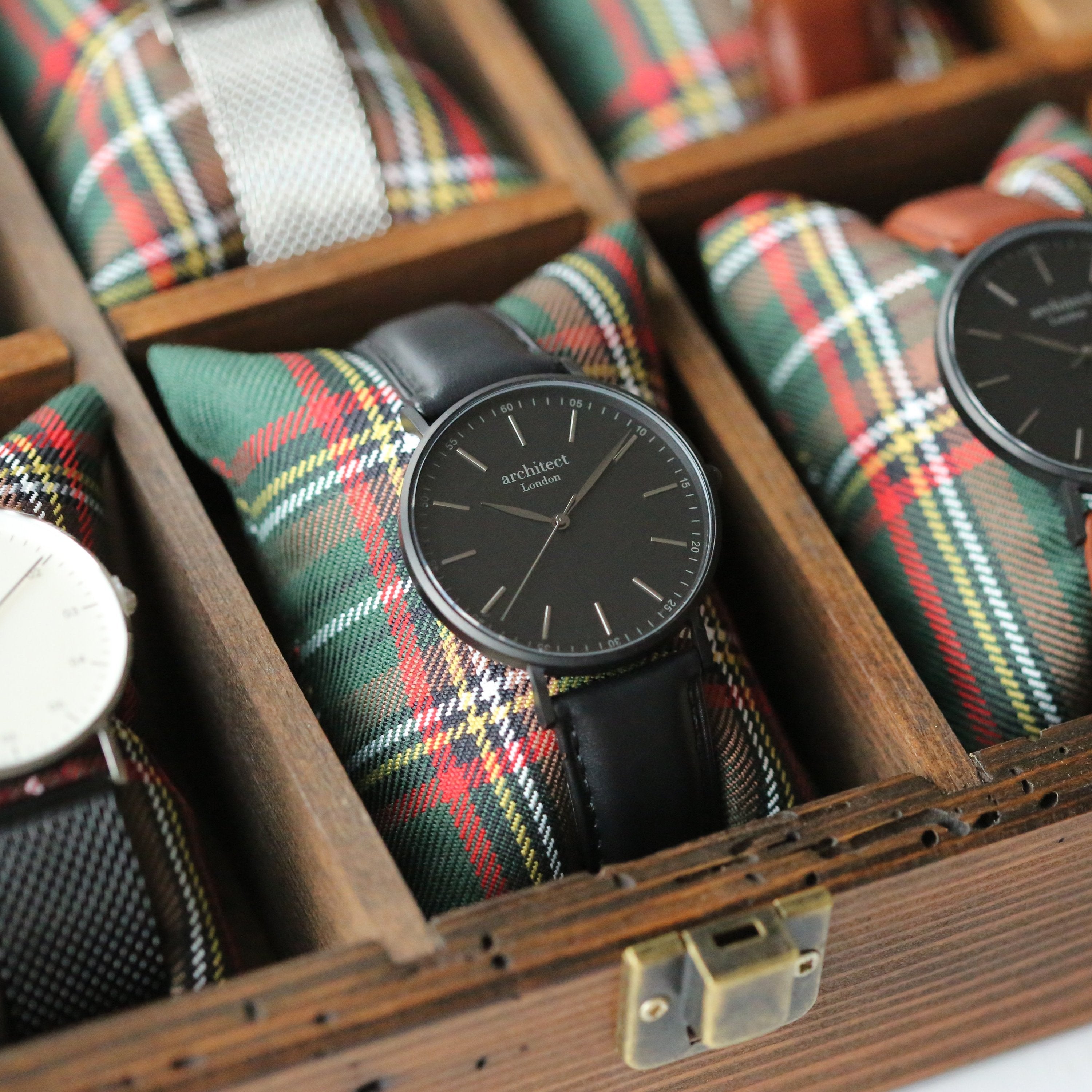 Watch with black strap and dark face displayed in a wooden box with plaid fabric